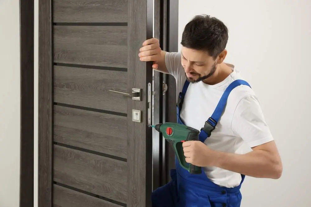 Worker In Uniform With Screw Gun Repairing Door Lock Indoors