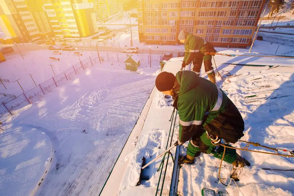 Snow Cleaning Team Of Male Workers Clean Roof Of Building From Snow With Shovels In Securing Belts Of Mantra