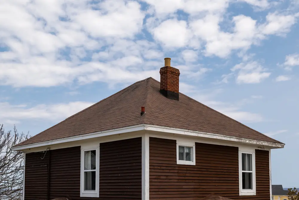 A Vintage Four Sided Brown Wooden House With An Asphalt Shingled Hip Roof There 039 S A Red Brick Chimney And Vent The Exterior Walls Are Brown Narrow Clapboard Siding With Multiple Small Glass Windows